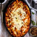 Bake rigatoni with pepper jack and mozzarella cheese, marinara sauce, topped with red pepper flakes in black dish.